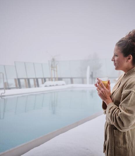 Frau im Bademantel trinkt Tee am verschneiten Infinty-Pool